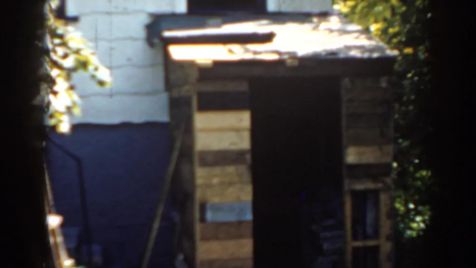 1963 Florida home-movie frame of a weathered backyard shed with uneven wooden planks, a dark open doorway, foliage, and painted wall sections.