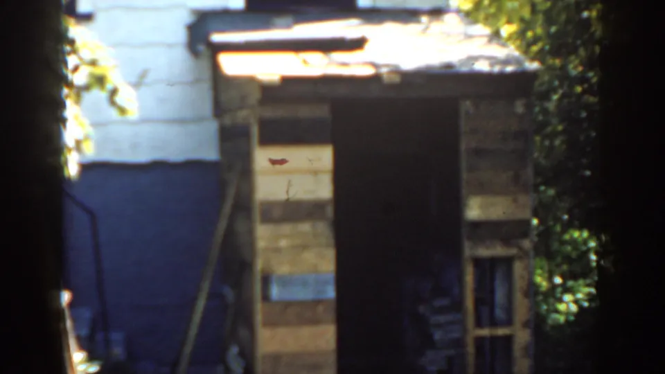1963 Florida home-movie frame centered on a rough backyard shed, showing plank boards, a dark entrance, small lower window, and bright foliage.