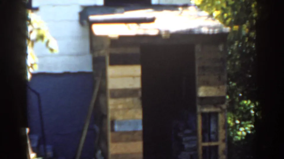 1963 Florida home-movie frame showing a patched wooden backyard shed, dark open doorway, small window, blue-and-white wall, and dense greenery.