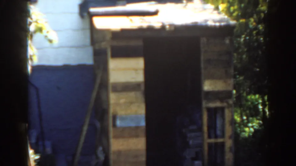 1963 Florida 8mm frame showing a rustic wooden shed near white siding, a blue lower wall, foliage, and strong backyard sunlight.