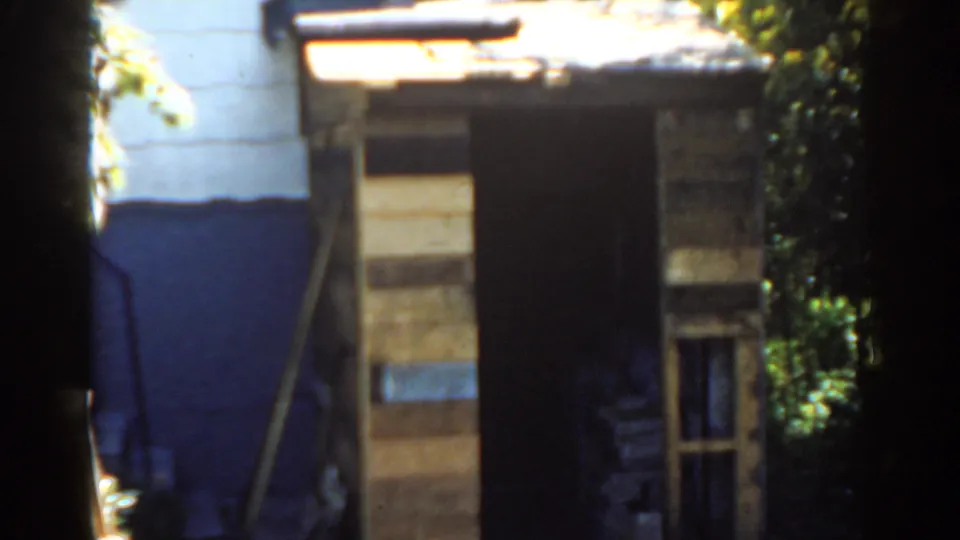 1963 Florida home-movie frame of a small wooden shed with a dark open doorway, painted house wall, and surrounding greenery.