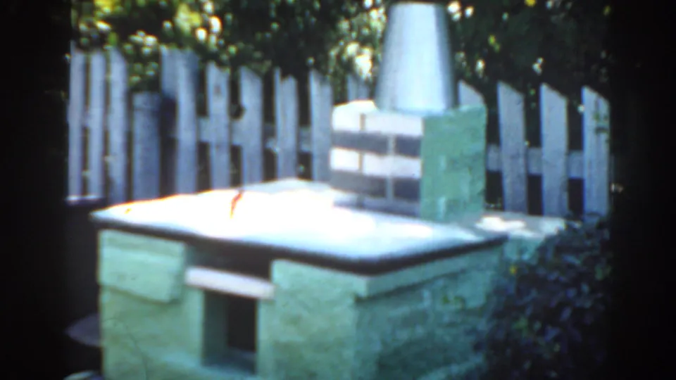 1963 Florida 8mm frame showing a low stone barbecue grill, brick chimney base, metal flue, white picket fence, and shaded greenery.