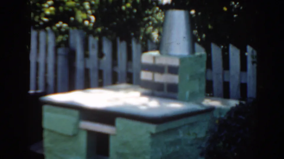 1963 Florida home-movie frame of a masonry backyard grill with a brick-and-metal chimney, white fence, and dense garden shade.