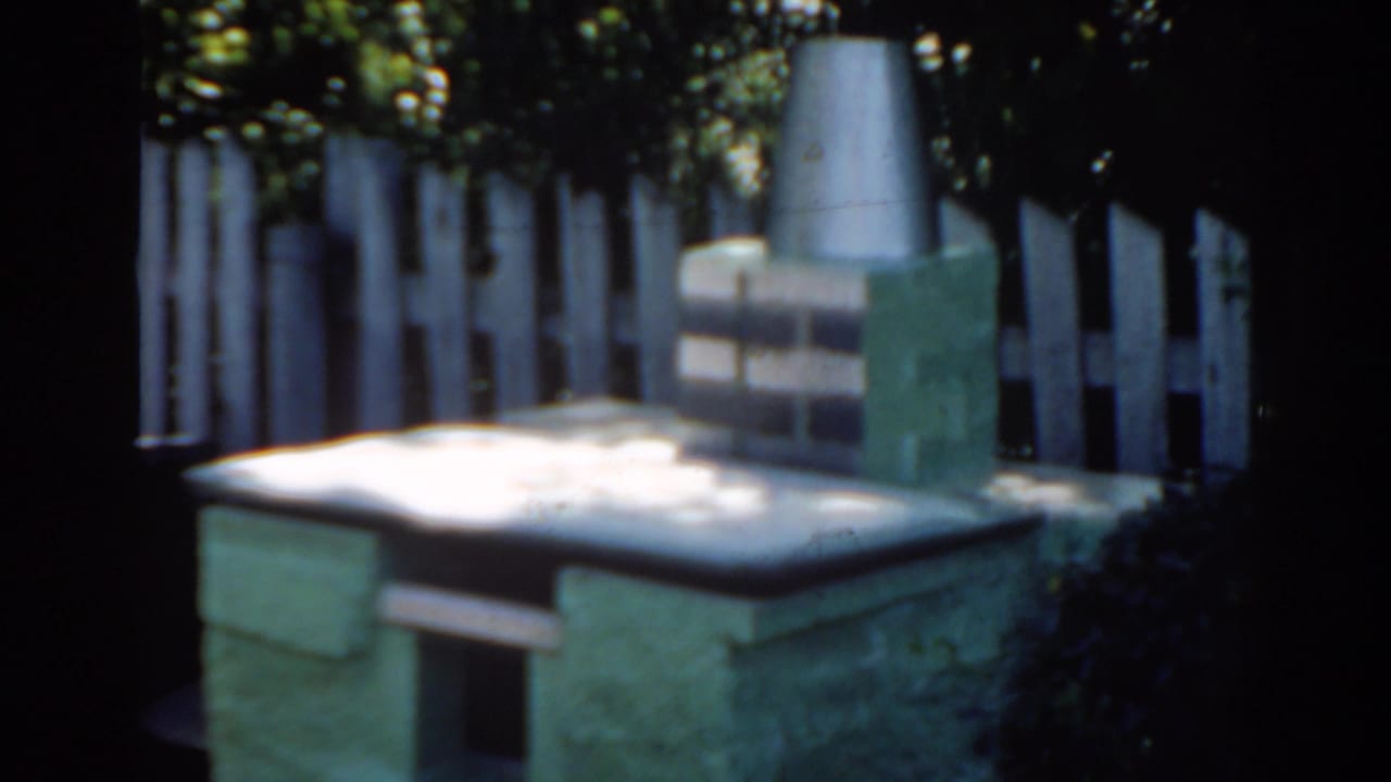 Small stone backyard structure with chimney beside a white picket fence.