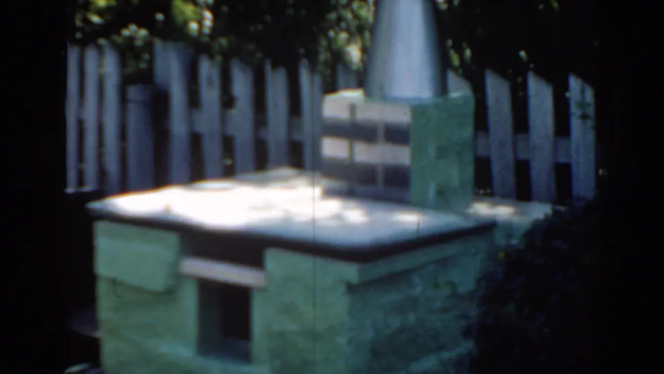 1963 Florida home-movie frame of a small stone backyard structure, possibly an outdoor oven or pet house, with fence and foliage.