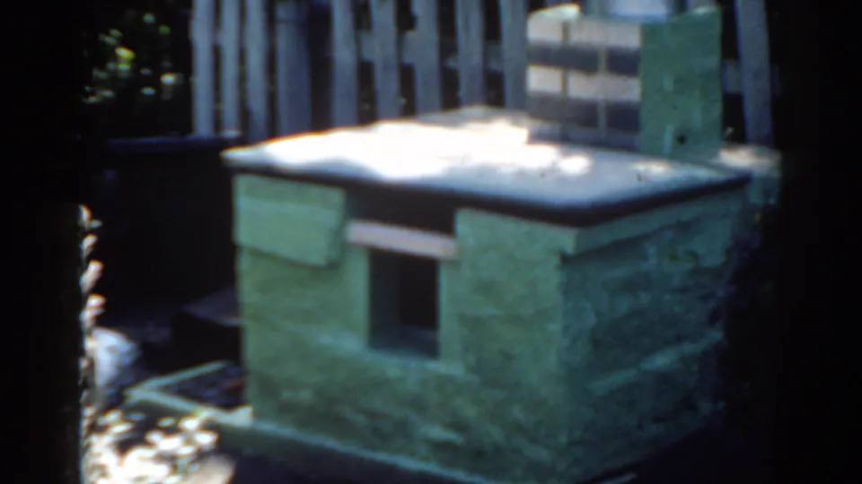 1963 Florida 8mm still of the masonry backyard object with a front opening, brick chimney, metal cap, fence, and plants.