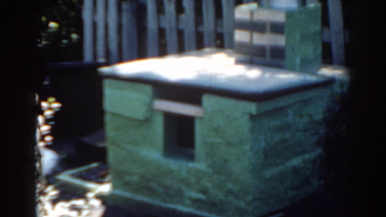 Green masonry outdoor structure with snow on top beside a fence and dark foliage.