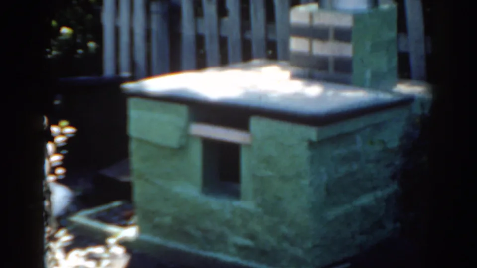 1963 Florida 8mm frame showing a green masonry outdoor structure capped with snow near a wooden fence and shaded foliage.