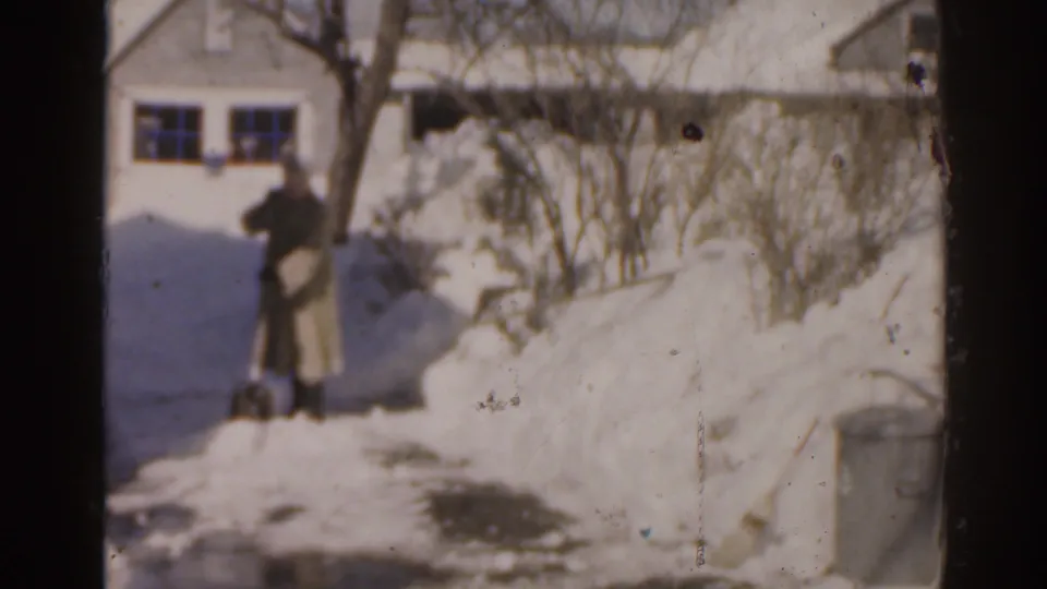 1963 Florida 8mm frame showing a bundled adult near snowbanks outside a residential house with bare shrubs and a walkway.