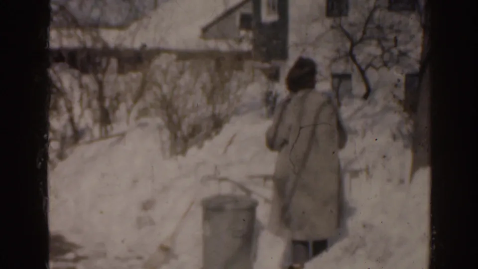 1963 Florida home-movie frame showing a bundled adult from behind near snowbanks, bare shrubs, and a residential house exterior.