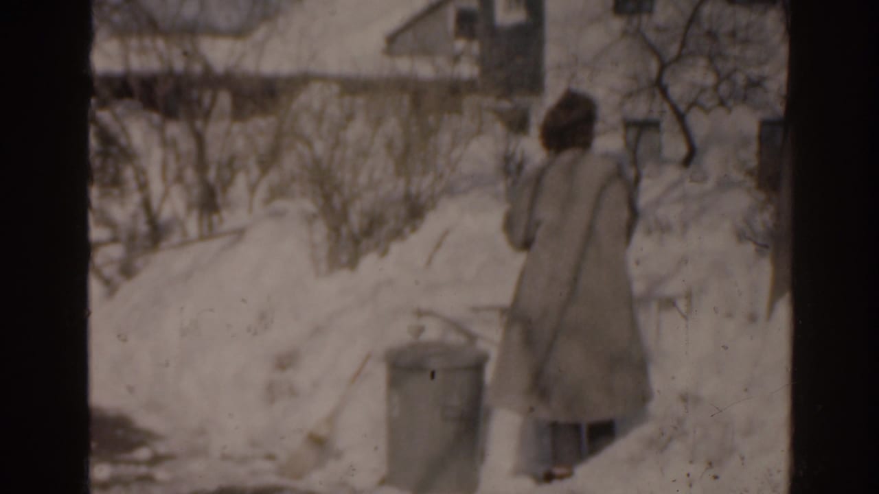 Bundled person walks through deep snow near houses and a trash can.