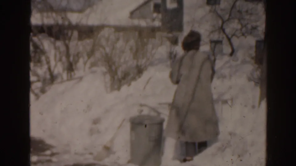 1963 Florida home-movie frame showing a bundled person moving through deep snow beside residential houses and bare shrubs.