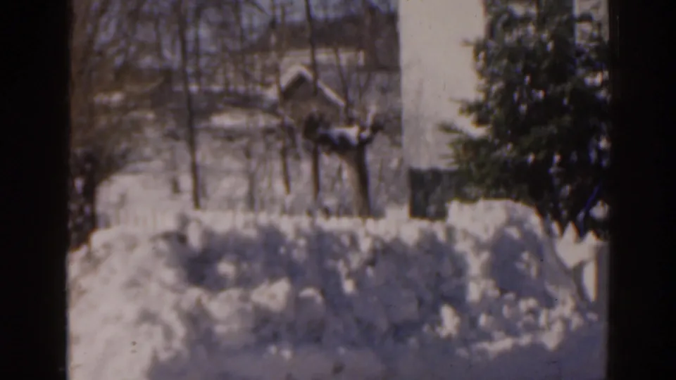 1963 Florida home-movie frame of a snow-filled yard with bare trees, a house wall, an evergreen, and piled snow.