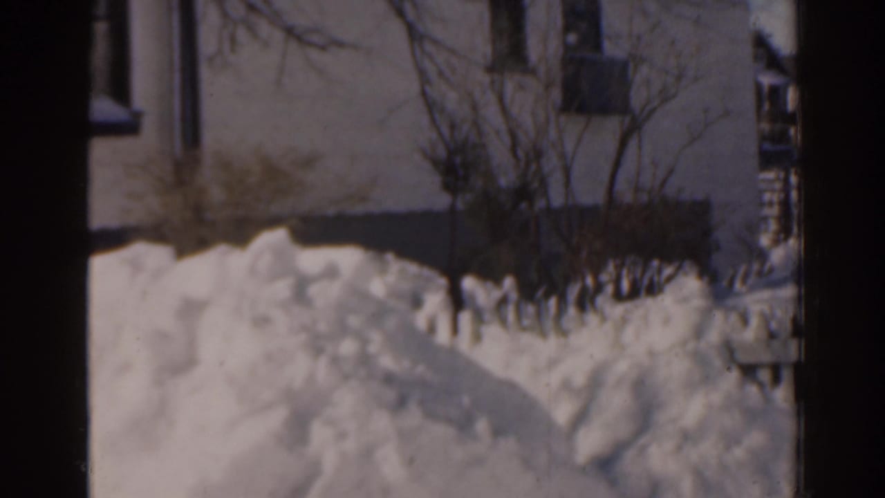 Snow piled beside a house exterior with shrubs and a partial window.