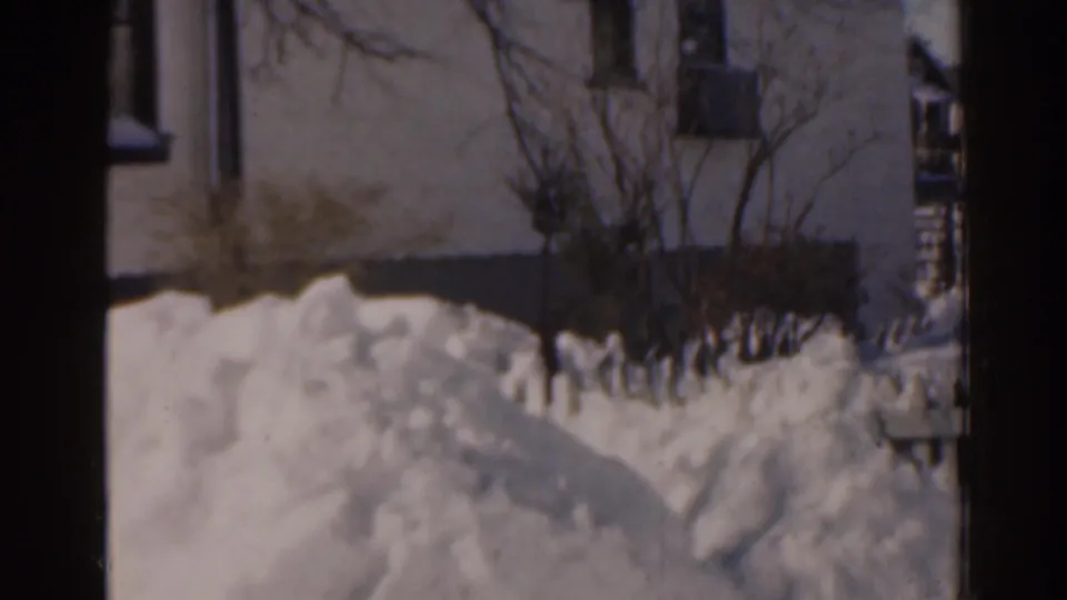 1963 Florida home-movie frame of snowbanks against a house exterior, bare shrubs, and a dark vertical film edge.