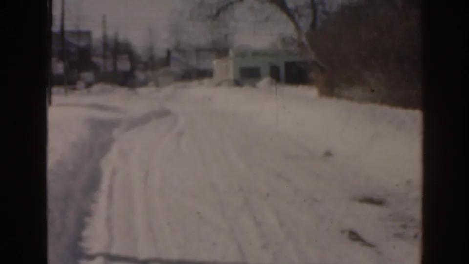 1963 Florida 8mm view down a snow-covered road with tire tracks, roadside snowbanks, bare trees, and distant structures.