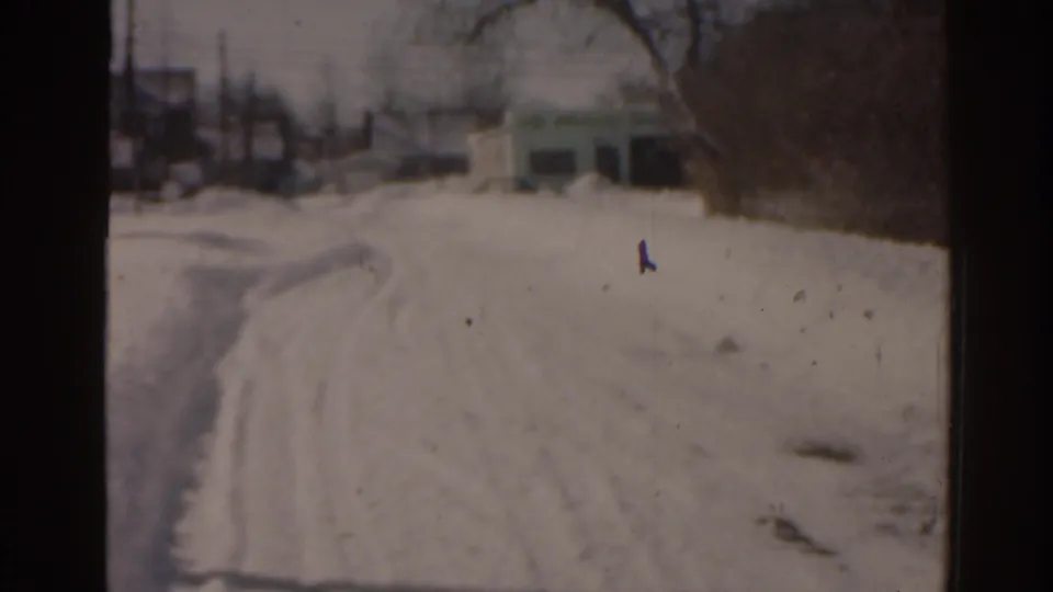 1963 Florida home-movie frame showing a snowy road, tire tracks, roadside snowbanks, bare trees, and distant structures.