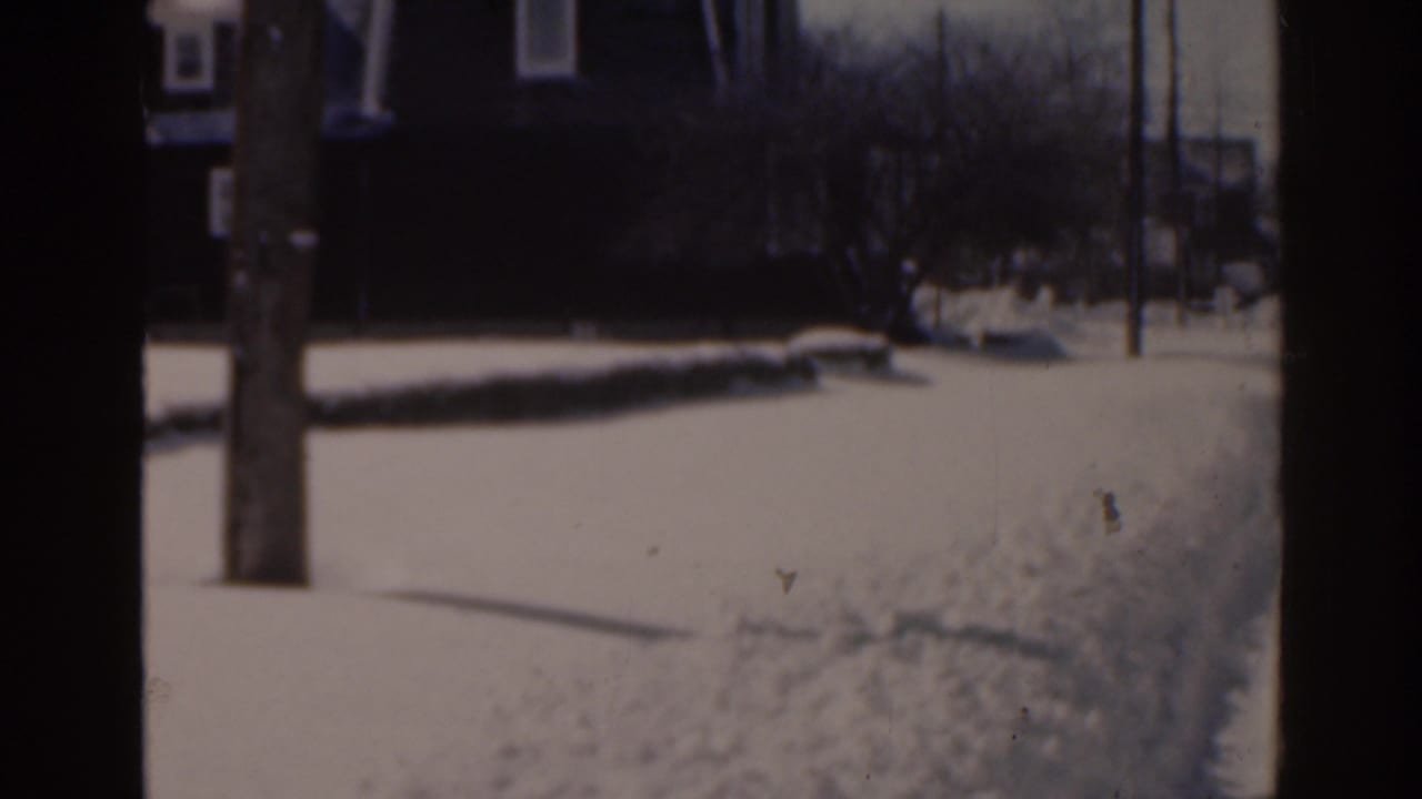 Snow-covered residential sidewalk with a utility pole and houses in the distance.
