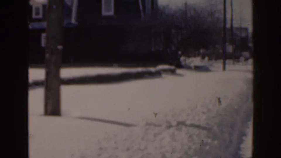 1963 Florida home-movie frame showing snow along a residential sidewalk, a wooden utility pole, bare trees, and distant houses.