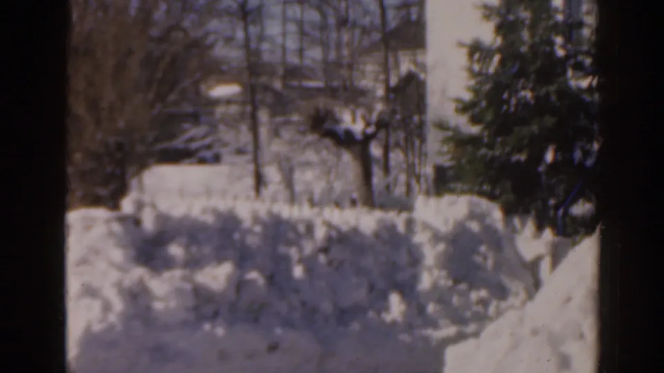 1963 Florida 8mm frame showing a snow-covered yard, bare trees, an evergreen shrub, and residential buildings in the background.
