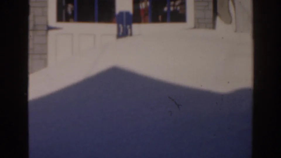 1963 8mm frame shifting to a sunlit exterior with a large shadow, building entrance, and distant people.
