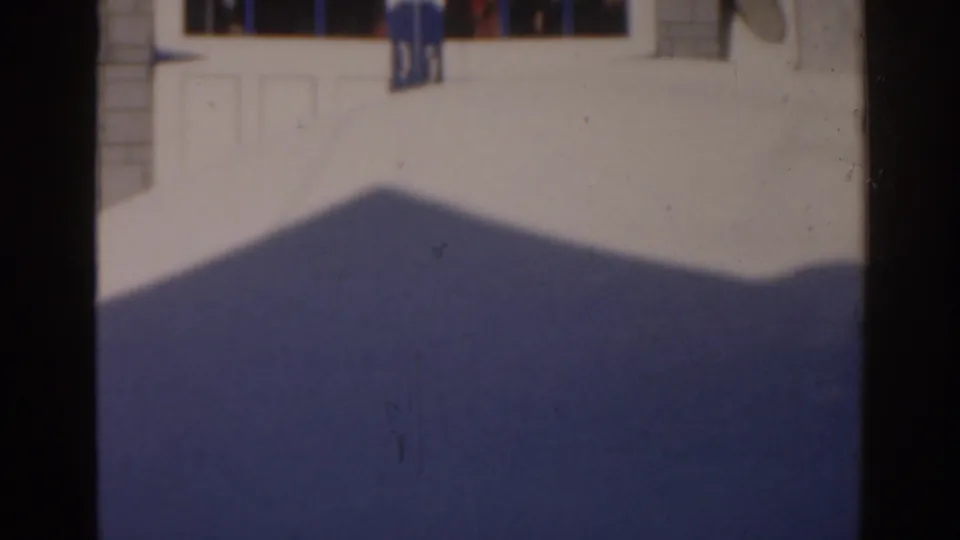 1963 8mm exterior view of a bright building area with distant people and a large shadow crossing the foreground.