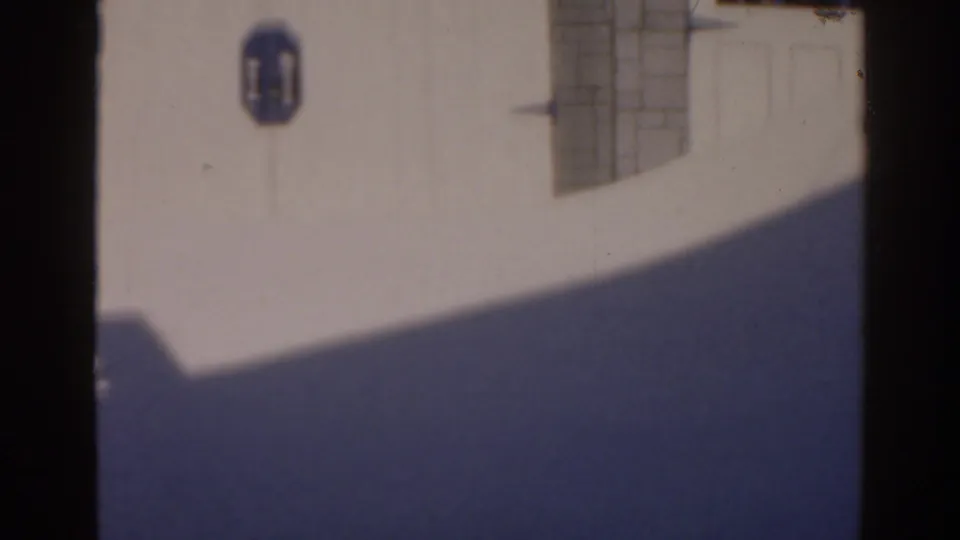 1963 Florida 8mm frame showing a white building wall with a blue octagonal sign, gridded window, and deep diagonal shadow.