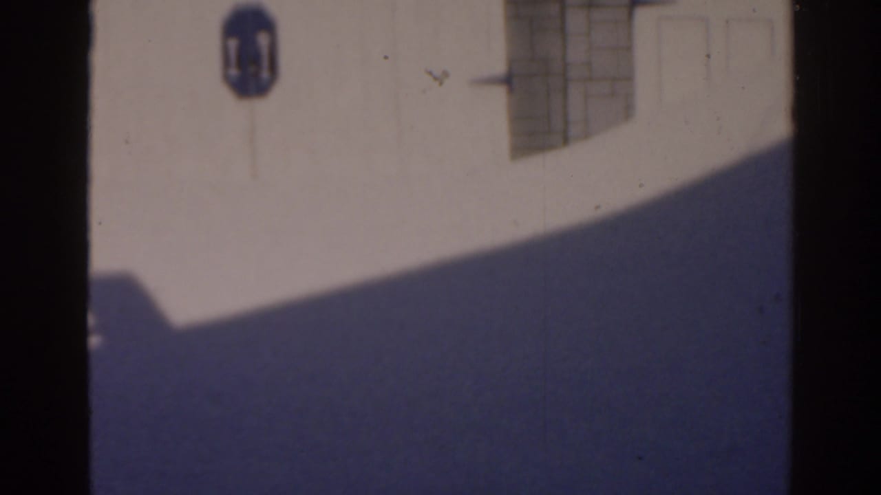 1963 Florida home-movie frame of a sunlit white wall, blue wall sign, gridded window, and broad blue-gray shadow across the facade.