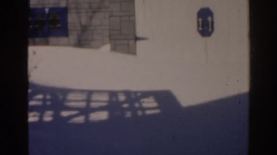 1963 Florida 8mm frame showing a white building facade with a blue sign, arched window grille, and dark metal fence shadows.