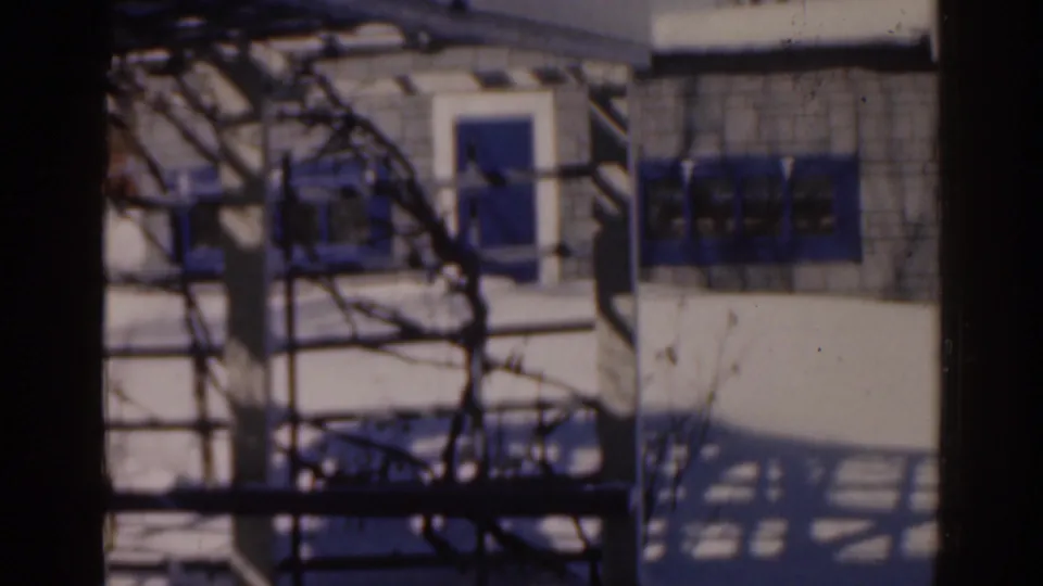1963 Florida home-movie frame showing a white exterior with a doorway, blue shaded windows, metal framework, and sharp fence shadows.