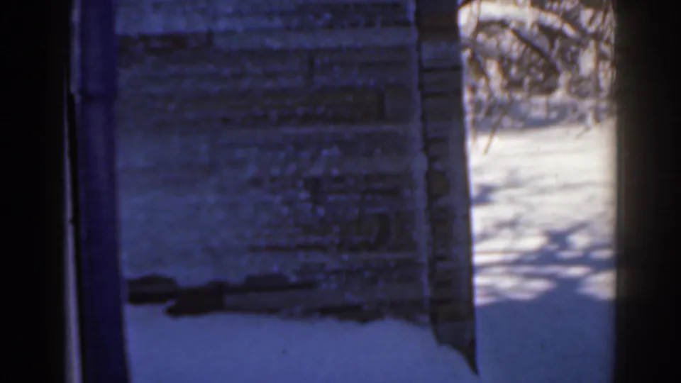 Blue-tinted 1963 8mm frame showing snow beside a textured rural wall, with bare branches and dark edge framing.