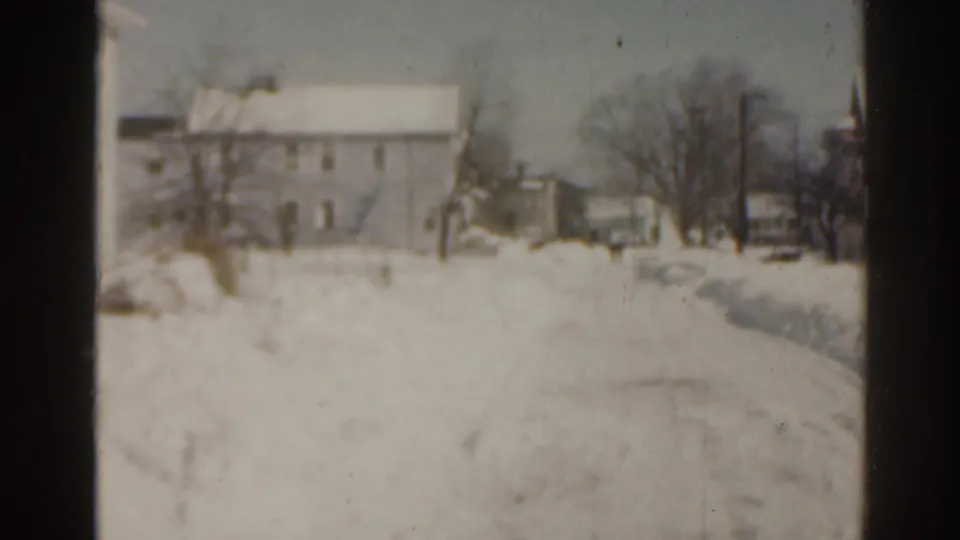 1963 Florida frame showing a snow-covered house beyond a snowy yard, with bare trees, poles, and a cleared or worn path visible.
