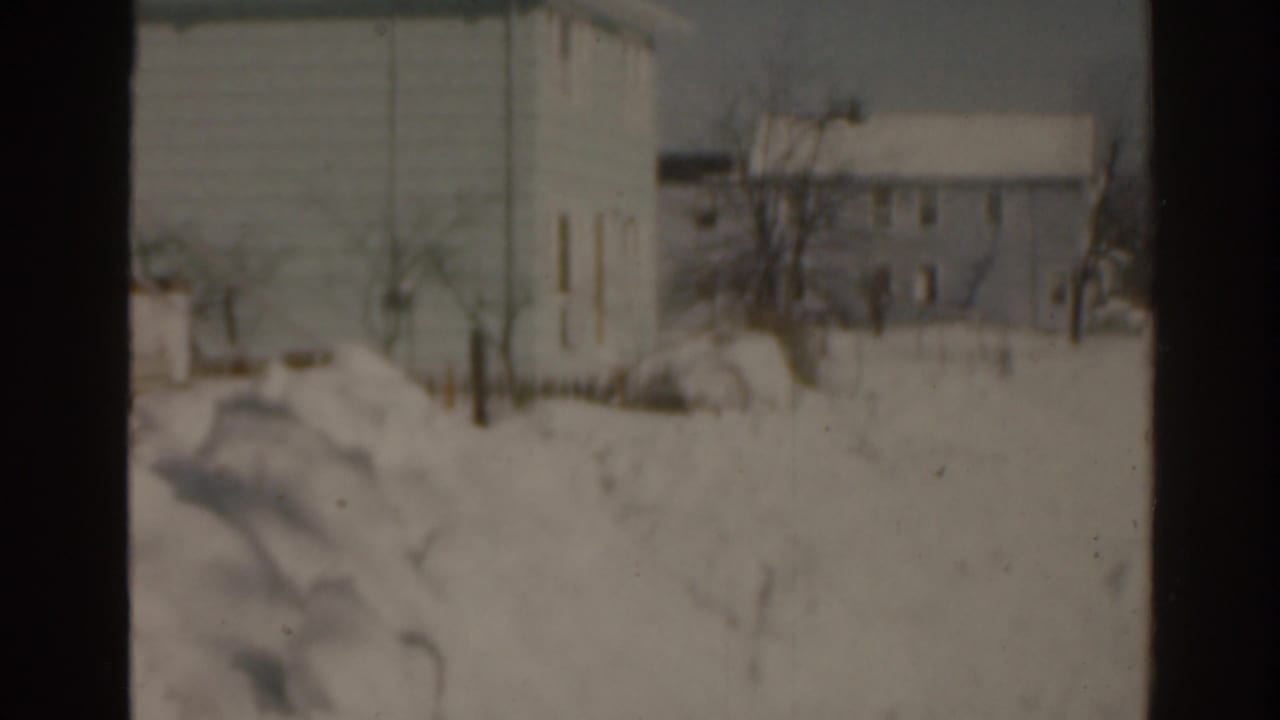 Snow-covered residential street with houses, bare trees, and a large pale building in the background.