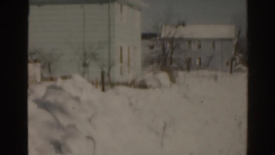 1963 Florida home-movie frame showing deep snow across a residential area, with houses, bare trees, and a larger building beyond.