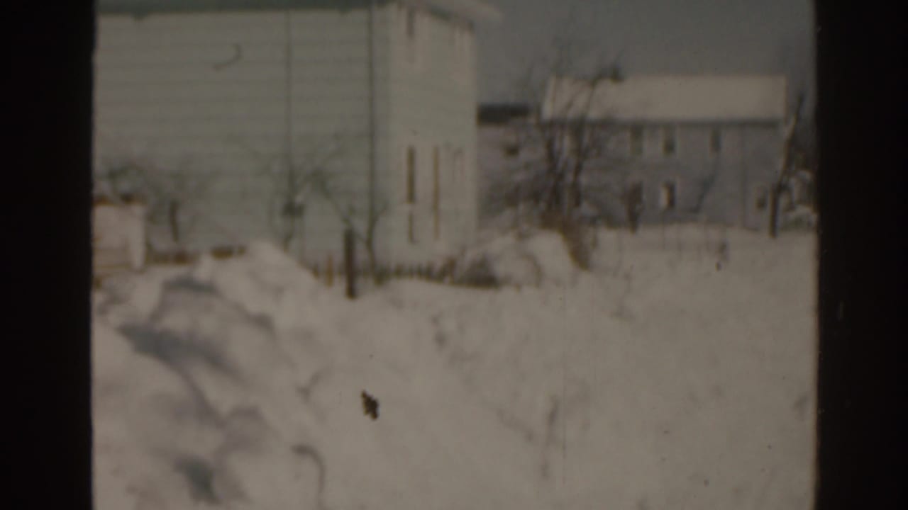 1963 Florida 8mm frame of a snow-covered residential neighborhood, with bare trees and houses seen in soft focus.