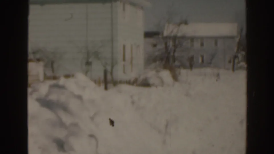 1963 Florida 8mm frame of a snow-covered residential neighborhood, with bare trees and houses seen in soft focus.