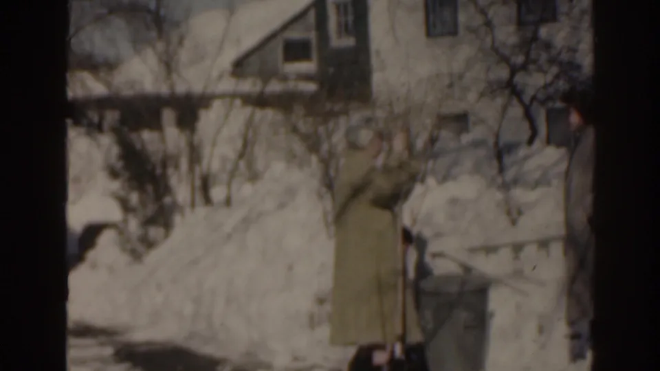 1963 Florida home-movie frame showing a bundled adult near deep snow, a trash can, bare branches, and nearby homes.