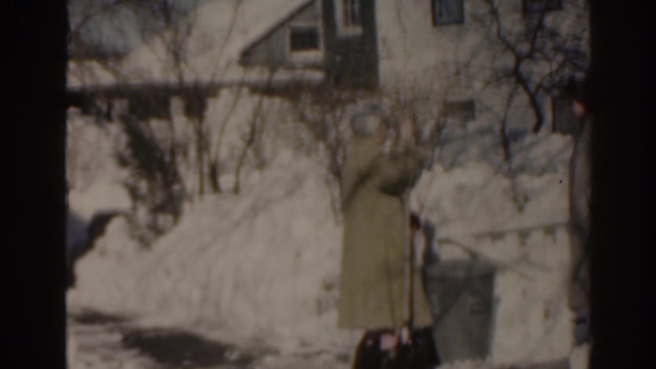 Two women in winter coats stand by a snowbank outside a house.