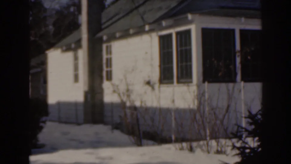 Residential house exterior in snow, with porch windows, bare shrubs, and a shaded yard in the 1963 Florida footage.