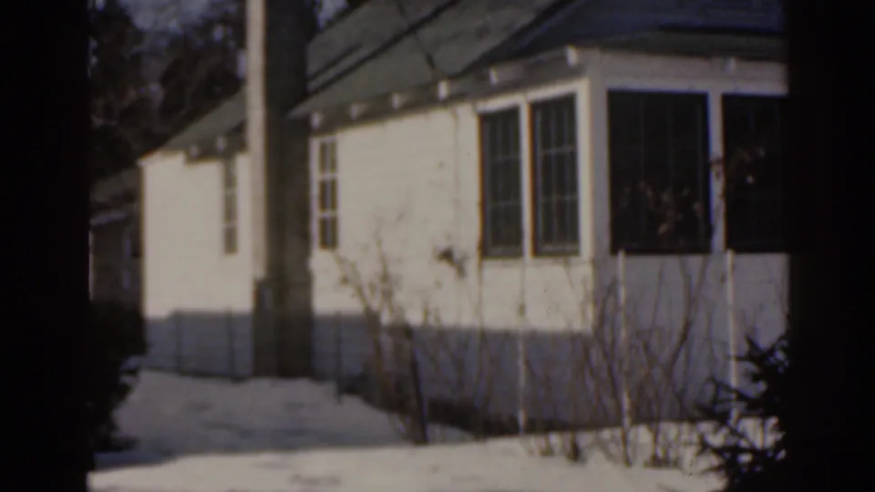 1963 Florida 8mm frame showing a white bungalow exterior with dark windows, tree trunks in front and pale ground cover around the yard.