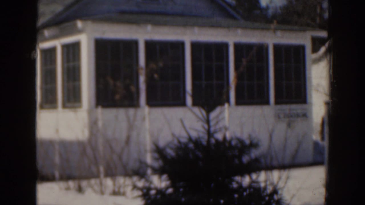 1963 Florida home-movie frame of a white house corner with large multi-pane windows, shrubs and a dark tree trunk crossing the foreground.