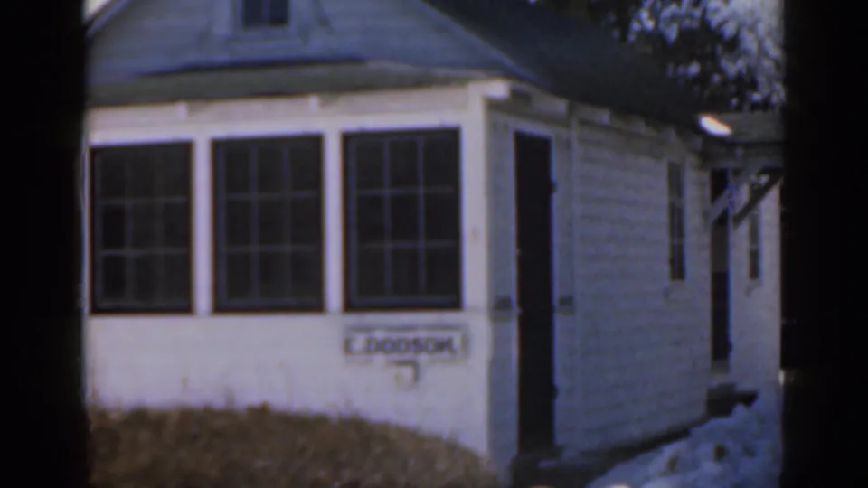 1963 Florida home-movie frame showing the side of the white bungalow, multi-pane windows, a doorway and the E. DOOSON nameplate.