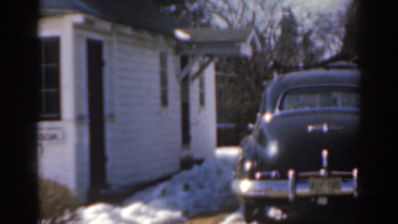 White house beside a parked dark sedan, with snow along the driveway.