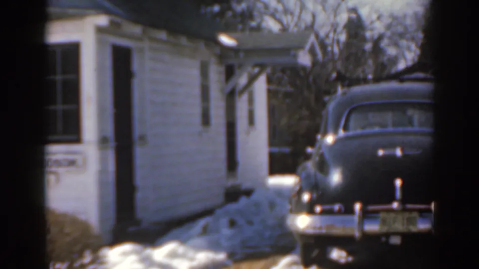 1963 Florida home-movie view of a white house wall, snowbank, and dark sedan with rear chrome details visible.
