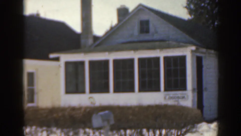 1963 8mm frame of a small white residential house with large front windows, hedges, bare trees, and snow on the ground.