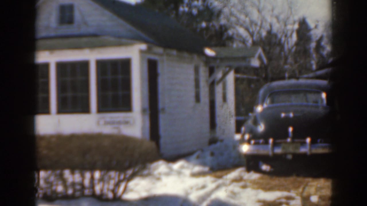 White house beside a dark vintage sedan parked near the driveway in snow.
