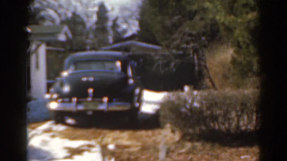 Rear view of a dark mid-century sedan with chrome details parked beside a residential house in a snowy 1963 8mm home-movie frame.