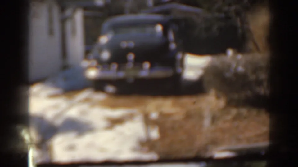 Soft 8mm frame of a dark sedan parked near a white house, with snow patches, a driveway, and winter shrubs visible.
