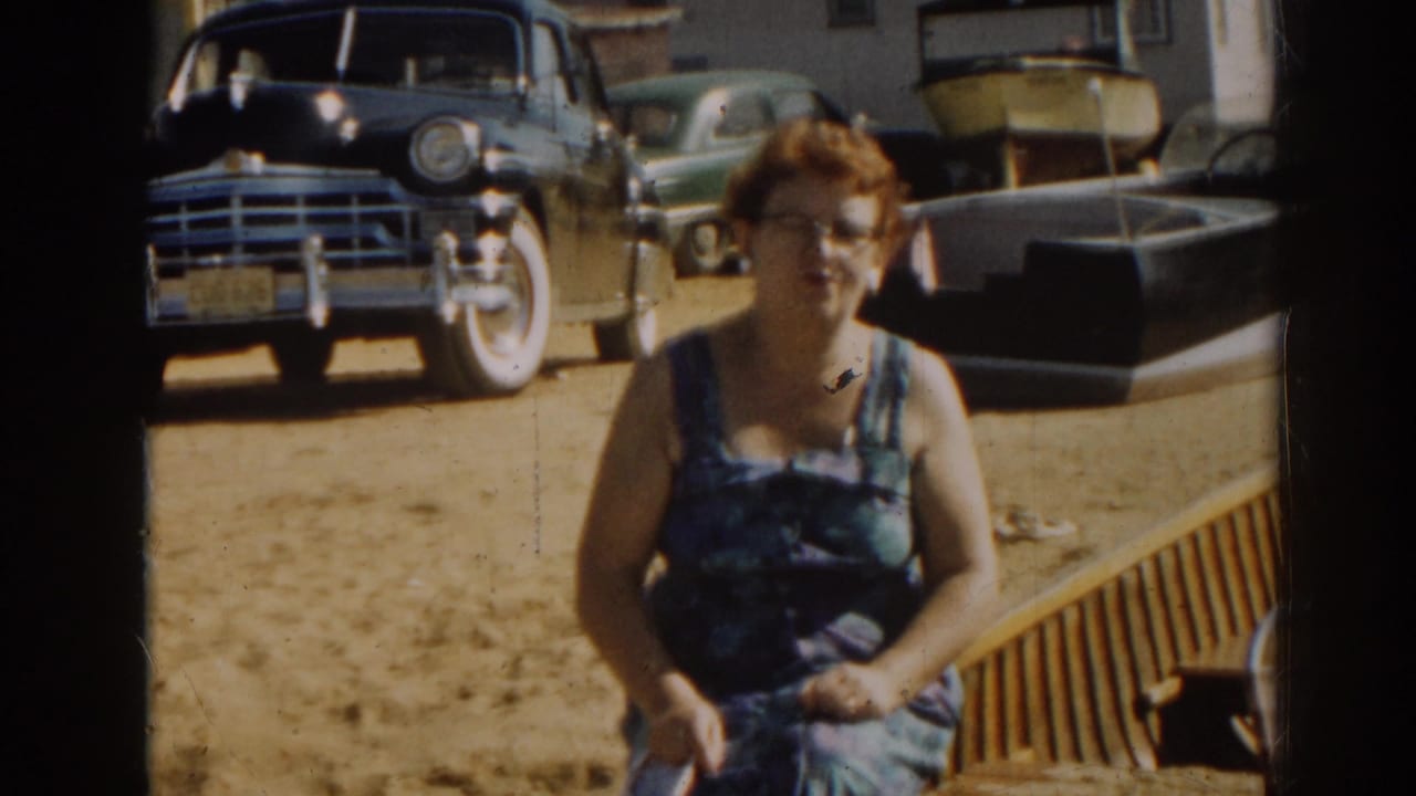 Woman in a blue patterned dress sits on sand with vintage cars and a boat behind her.