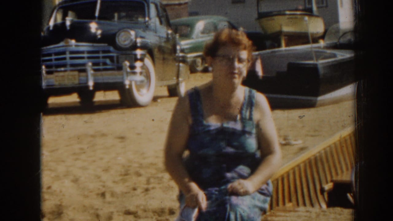 Seated woman in blue dress faces the camera near parked cars and small boats.